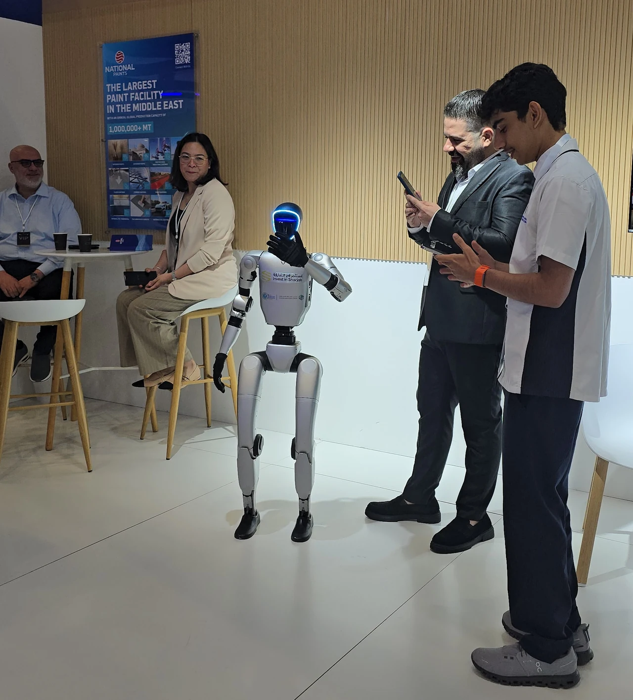 A humanoid robot beside an operator holding a controller and visitors at an indoor demonstration