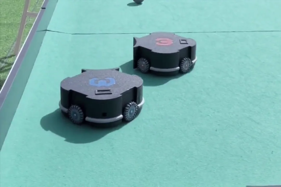 Two compact robots on the green playing surface of a robot-football arena