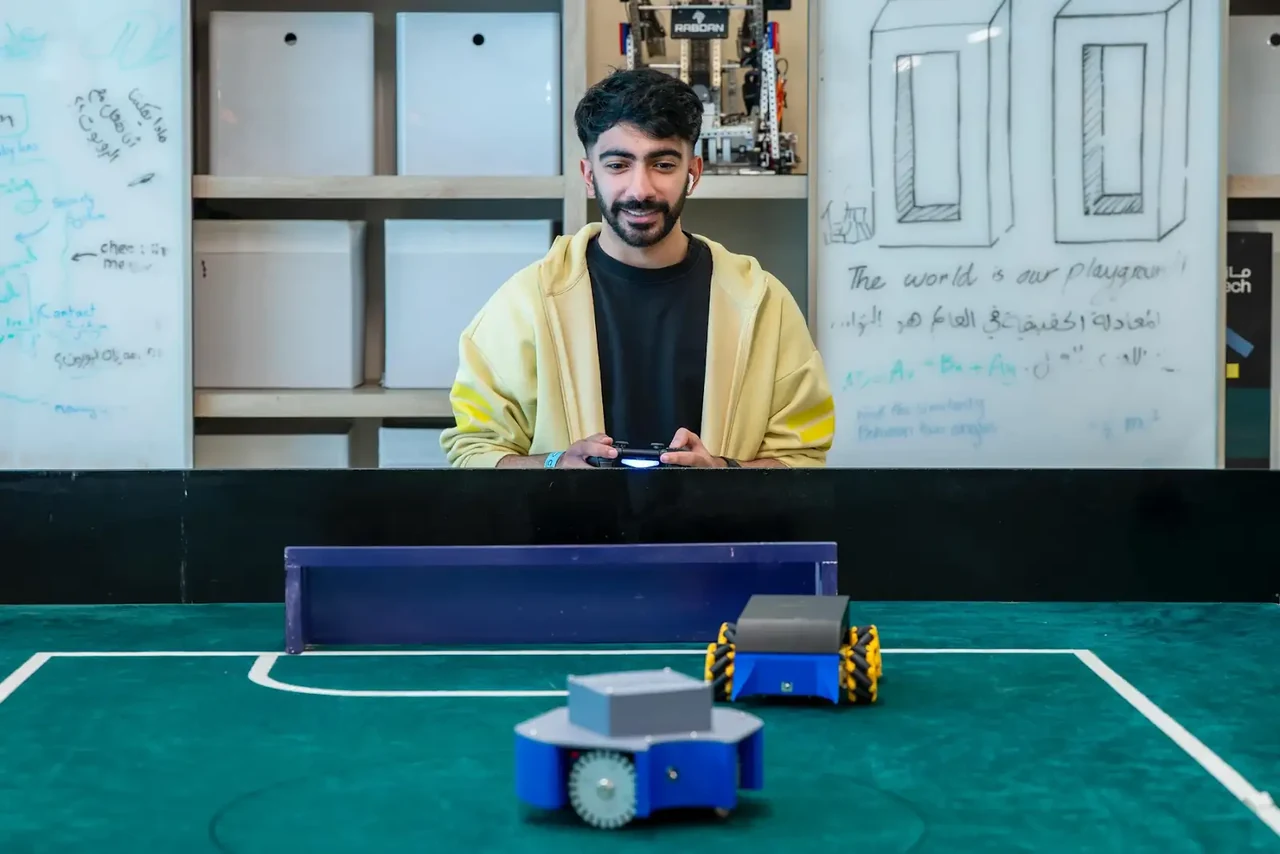 A participant holding a gamepad while robots play in the MazTech football arena